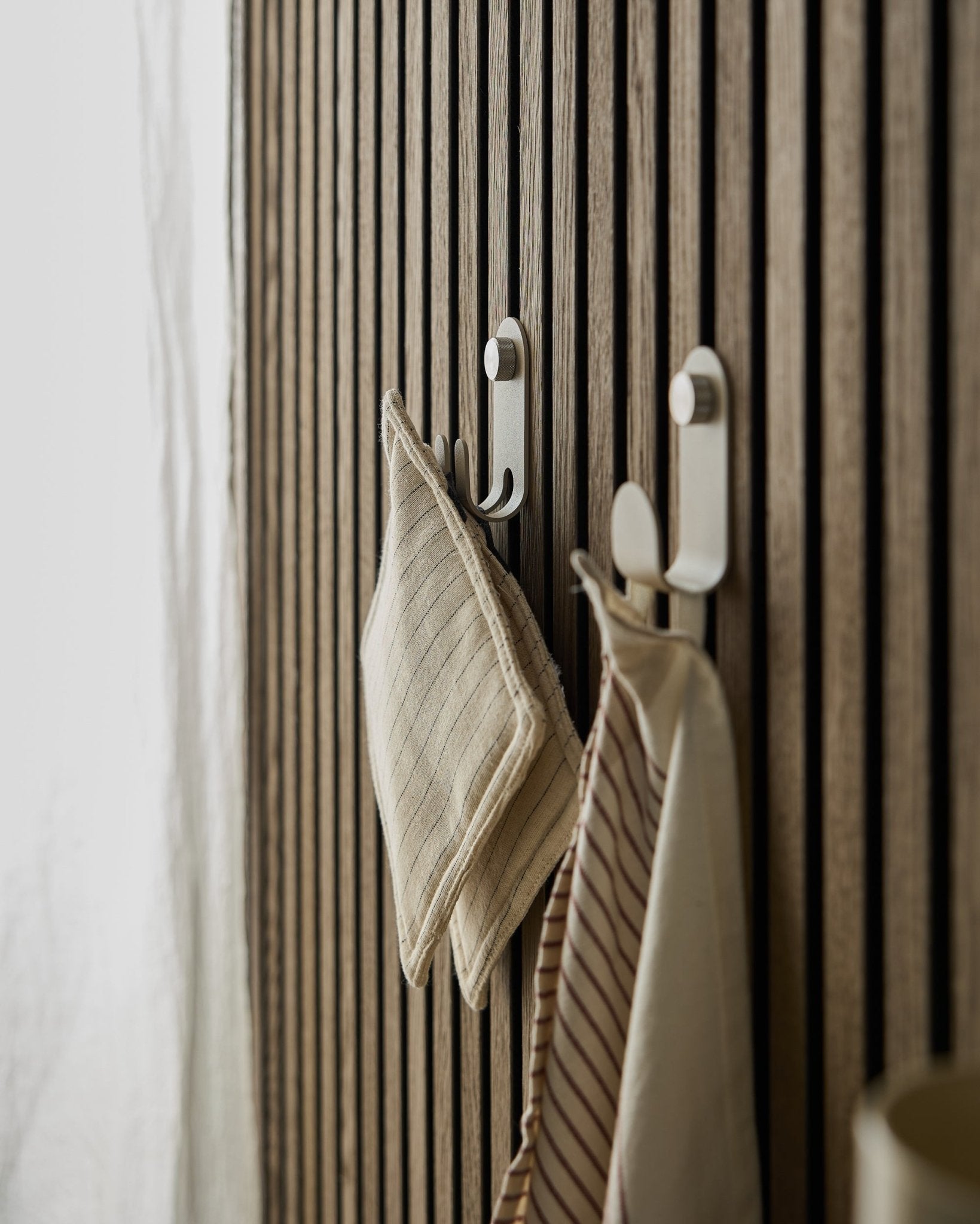 Due strofinacci a righe sono appesi al Norr Double Hanger di WoodUpp, montato su una slatwall nordica beige Pannello acustico, illuminata da una morbida luce naturale.